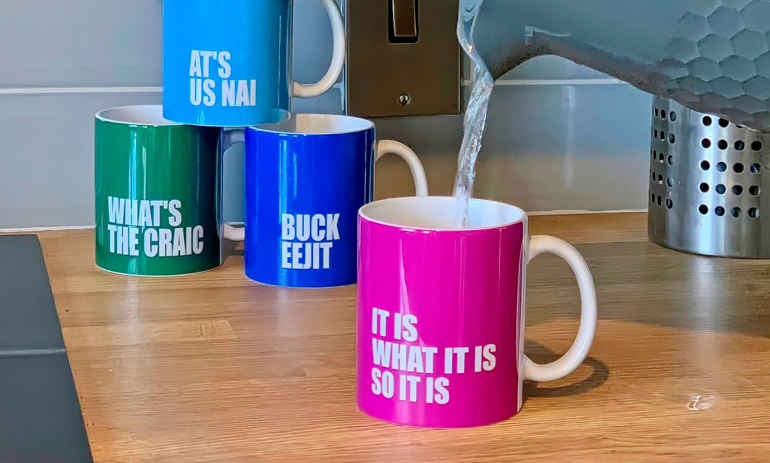 Photograph of the Northern Irish Slang Mugs in a kitchen with water being poured into the It Is What It Is Mug.